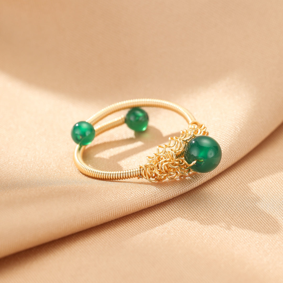 Bague filigrane dorée avec trois petites perles d'agate verte disposées le long de l'anneau, sur fond beige satiné.