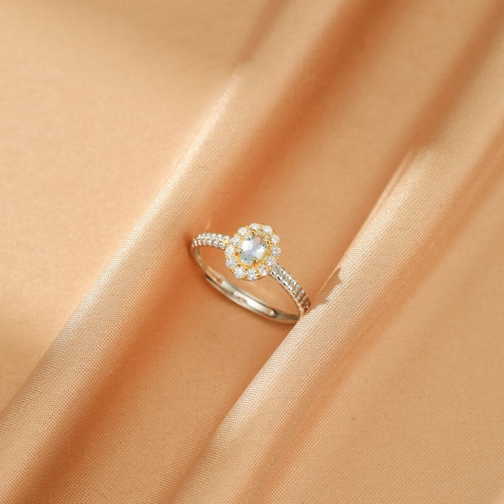 Bague en argent 925 avec une aigue-marine ovale entourée de petits cristaux scintillants, posée sur un tissu satiné couleur champagne.