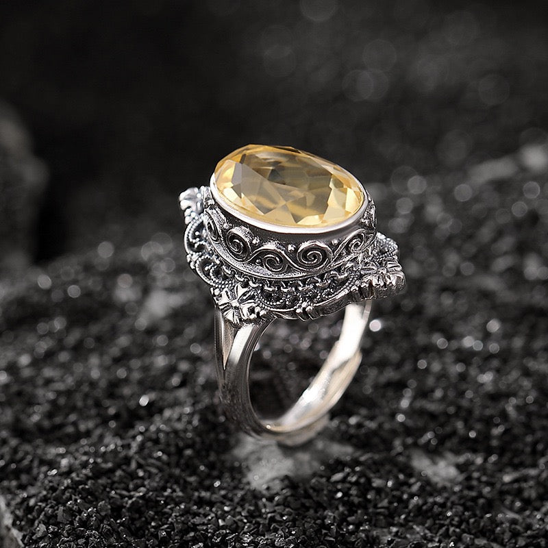 Bague élégante en argent ornée d'une grande citrine ovale, avec des détails finement ciselés, posée sur un fond sombre et texturé.