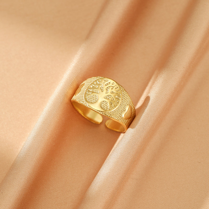 Bague ajustable dorée ornée d'un arbre de vie stylisé avec des motifs texturés sur le pourtour, posée sur un tissu satiné beige.