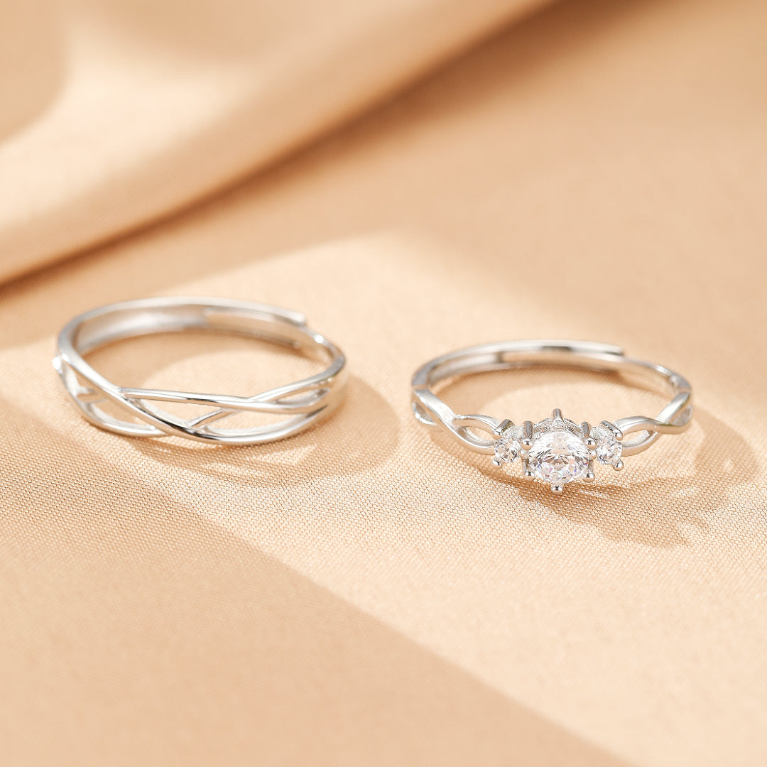 Deux bagues de couple en argent, l'une avec un design entrelacé simple et l'autre ornée d'une pierre centrale étincelante entourée de petites pierres.