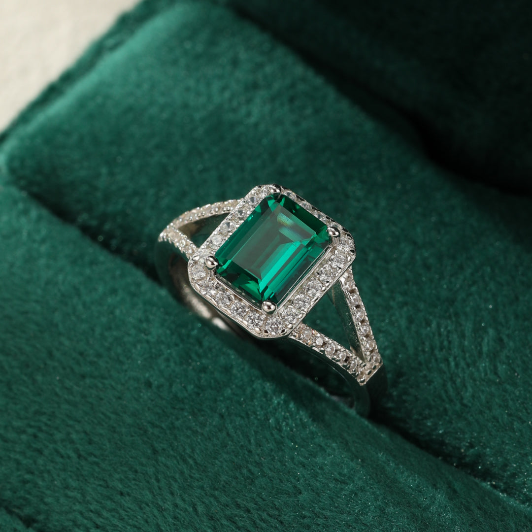 Bague en argent 925 avec une émeraude rectangulaire centrale entourée de petits diamants scintillants, posée sur un écrin en velours vert.