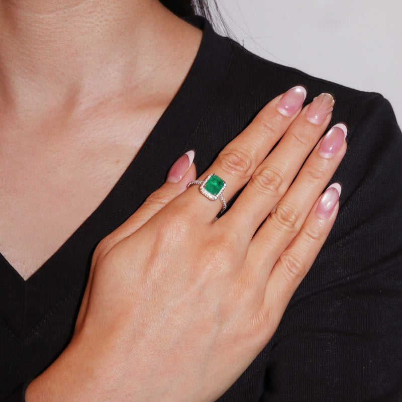 Bague en argent sertie d'une émeraude taille émeraude entourée de diamants, portée au doigt par une femme.