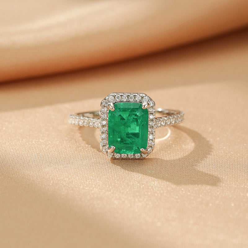 Bague en argent sertie d'une émeraude rectangulaire entourée de petits diamants scintillants, posée sur un fond de tissu satiné beige.