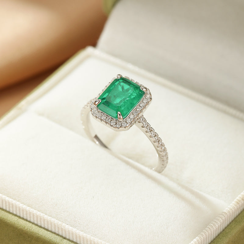 Bague en argent avec une émeraude rectangulaire taille émeraude entourée de petits diamants, posée dans une boîte à bijoux.