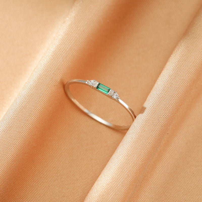 Bague fine en argent 925 avec une émeraude rectangulaire centrale et de petits diamants de chaque côté, posée sur un tissu satiné couleur pêche.