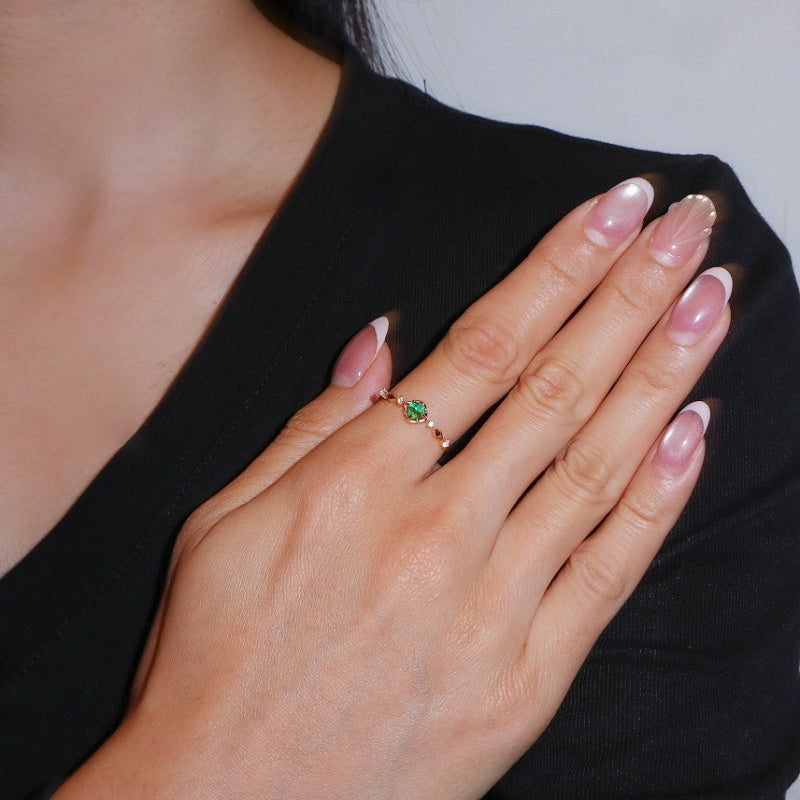 Bague Émeraude – Éclat Précieux en Argent Plaqué Or 14K, ornée d'une émeraude centrale et de petites pierres scintillantes sur la monture, portée au doigt d'une femme avec manucure française.