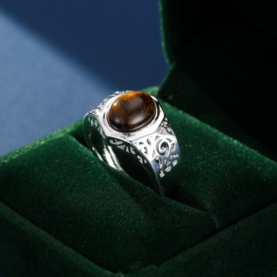 Bague homme argentée avec une pierre d'œil de tigre ovale et des motifs gravés sur le corps de la bague, présentée dans un écrin vert