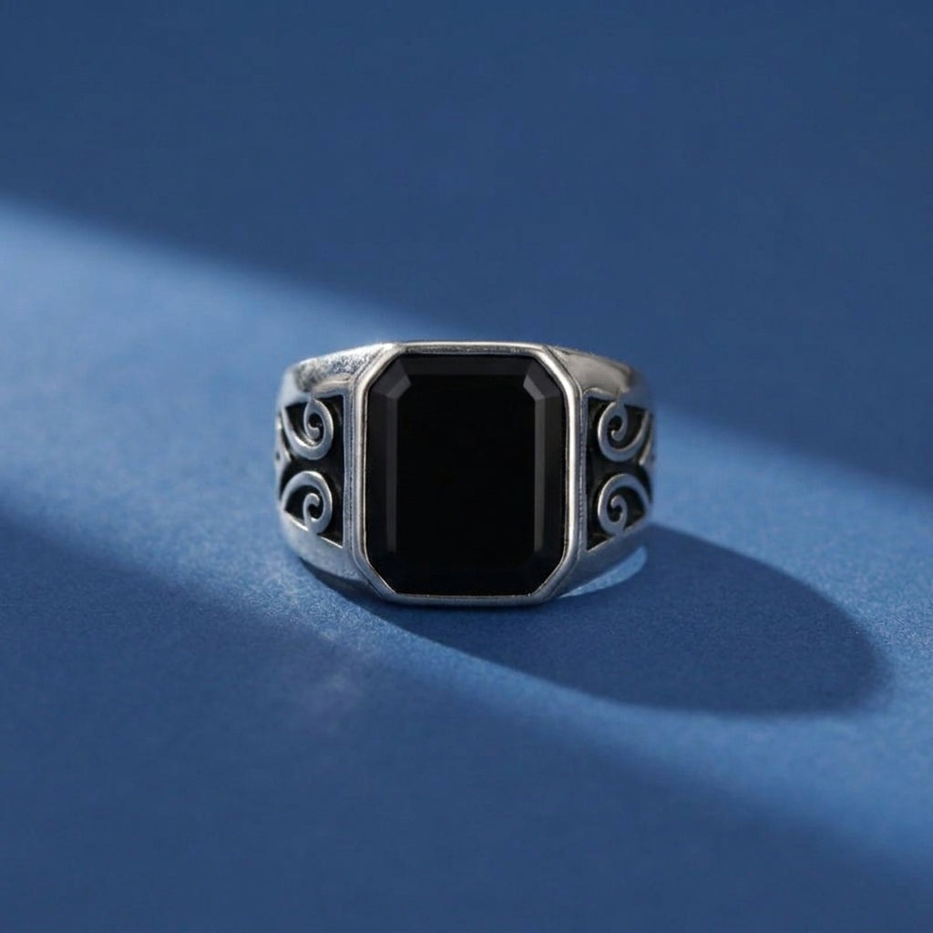 Bague pour homme en argent avec une pierre d'onyx noire rectangulaire et des détails gravés sur les côtés, posée sur un fond bleu.