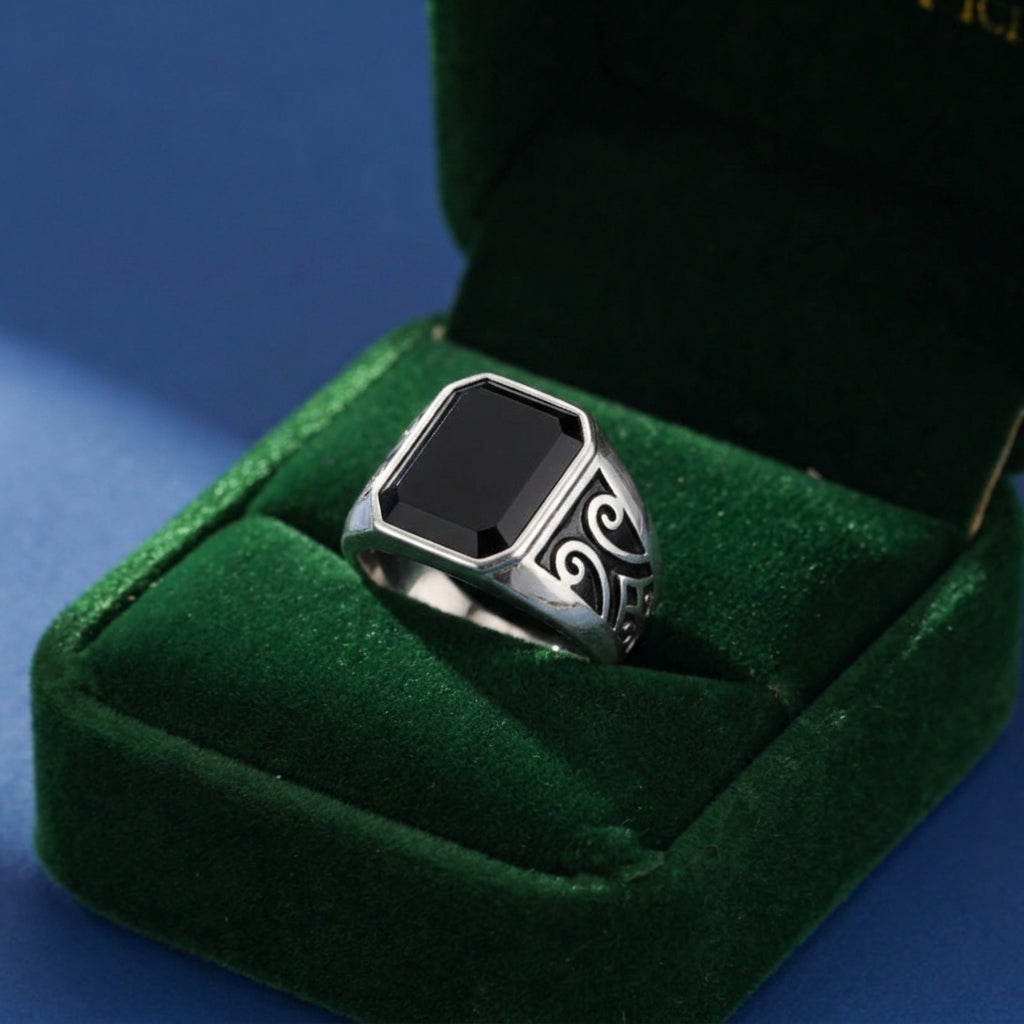 Bague pour homme avec pierre onyx noire rectangulaire et détails gravés sur les côtés, présentée dans un écrin en velours vert.
