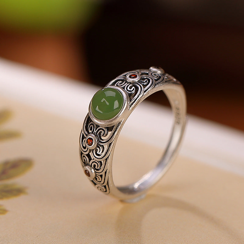 Bague en argent plaqué or avec un jade vert central et de petites pierres rouges sur les côtés, ornée de motifs sculptés.