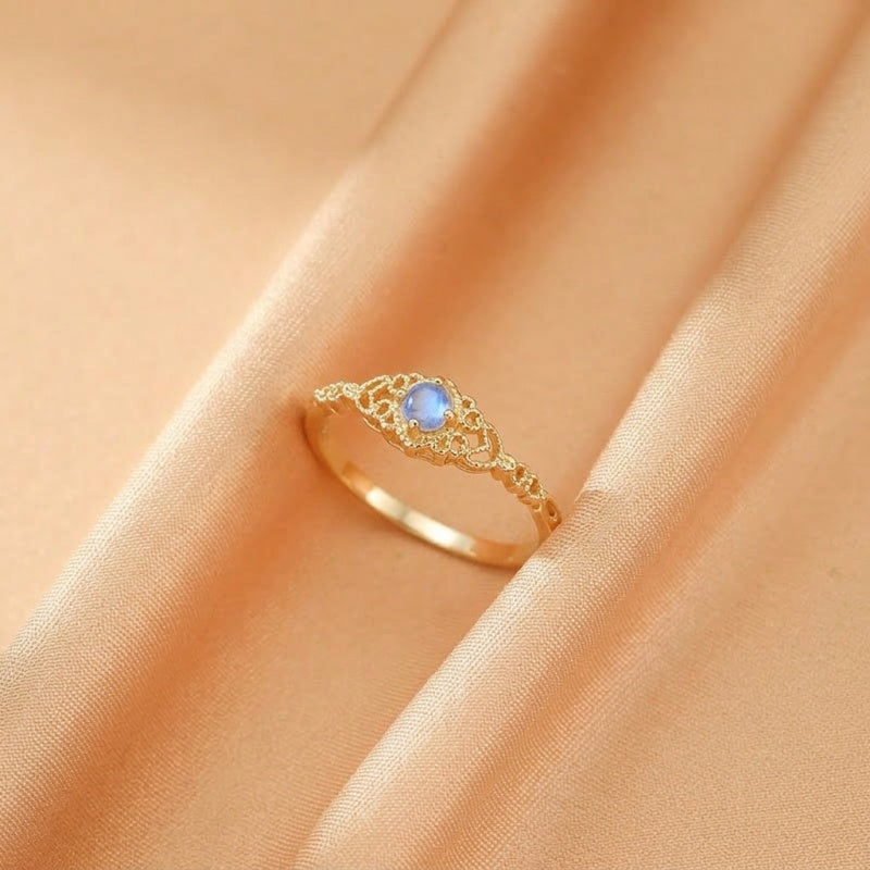 Bague dorée avec une pierre bleue centrale et des motifs ajourés sur le corps de la bague, posée sur un tissu satiné couleur pêche.
