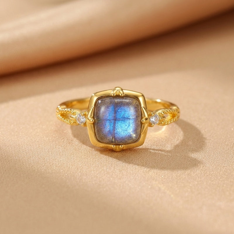 Bague en argent plaqué or sertie d'une labradorite bleue carrée et de deux petits diamants sur les côtés, sur un fond beige.