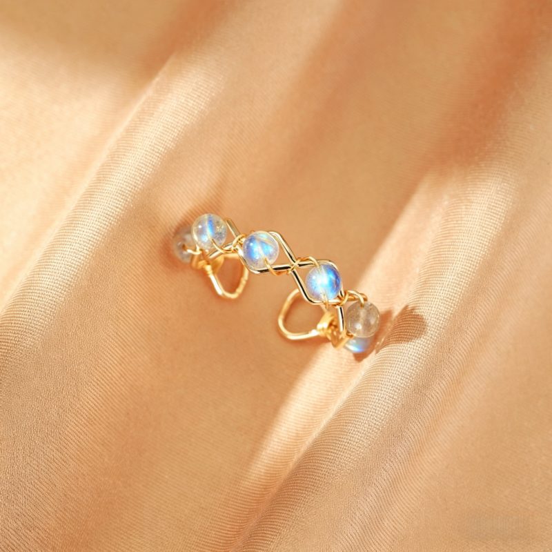 Bague ajustée en fil doré torsadé avec des petites perles de labradorite bleues iridescentes et grises, posée sur un tissu satiné pêche.