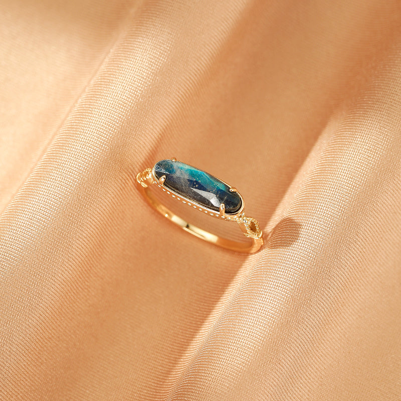 Bague en argent plaqué or avec une pierre de labradorite ovale aux reflets bleus et verts, mise en valeur sur un fond de satin couleur pêche.