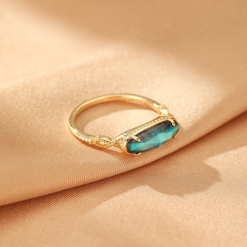Bague en argent plaqué or sertie d'une pierre de labradorite rectangulaire aux reflets bleus et verts, sur un tissu satiné doré.
