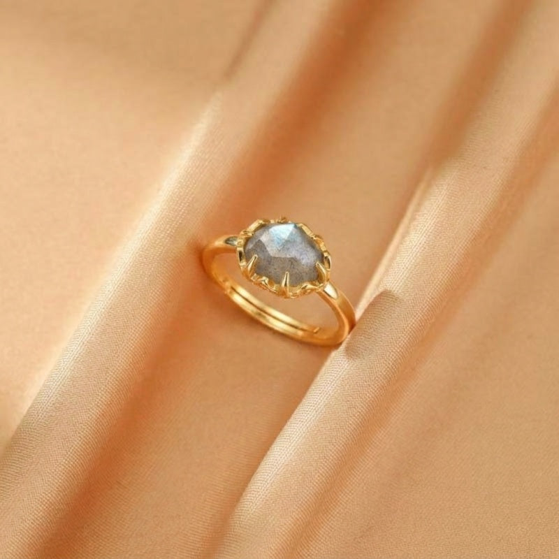 Bague en argent plaqué or sertie d'une pierre de labradorite grise aux reflets bleutés, sur un fond satiné doré.