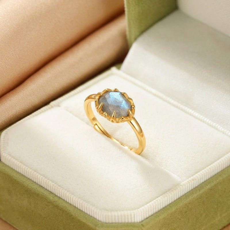 Bague en argent plaqué or avec une pierre de labradorite ovale grise aux reflets bleutés, montée sur un anneau ajustable et présentée dans un écrin vert.