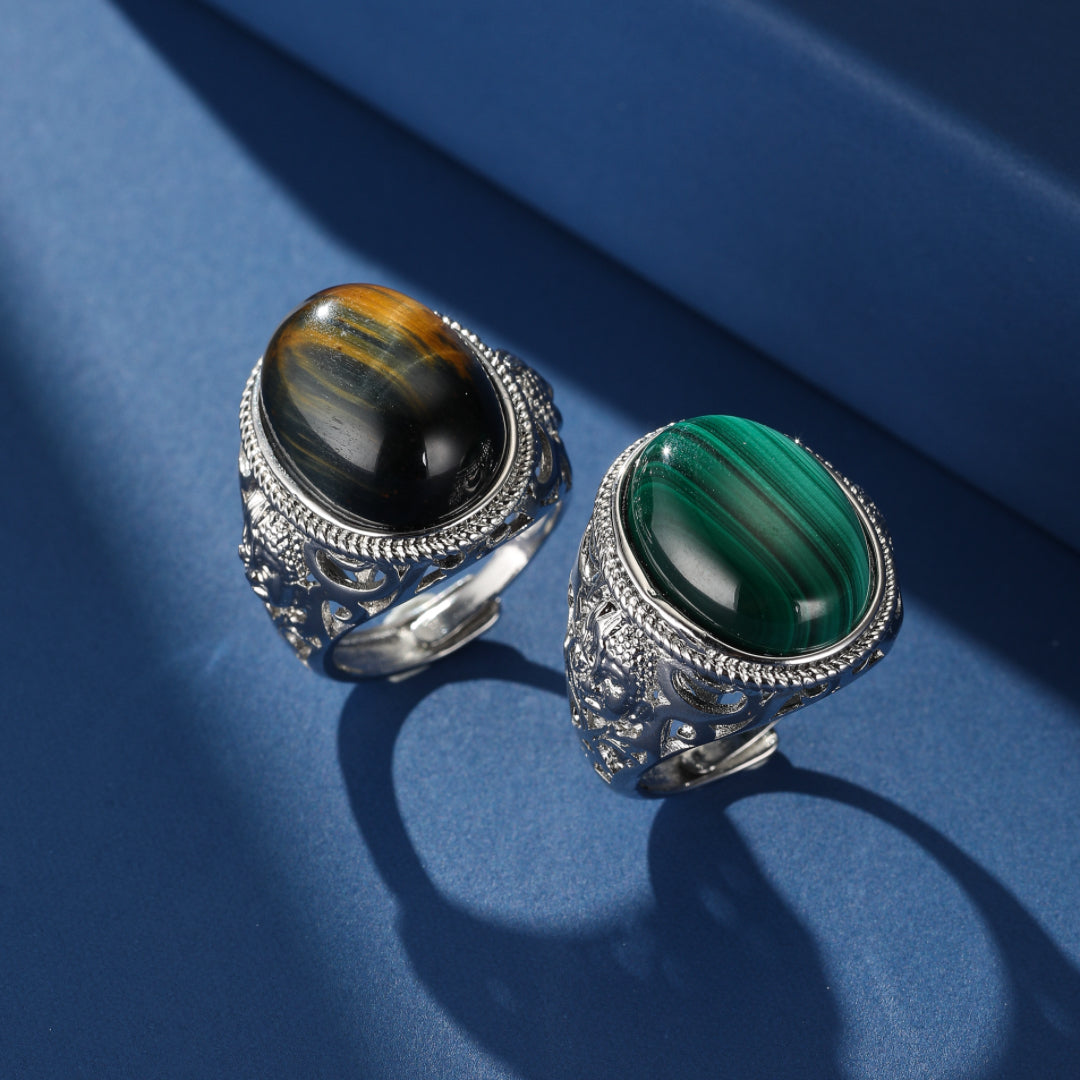 Deux bagues ajustables en argent 925, l'une ornée d'une pierre d'œil de tigre aux reflets noirs et dorés, l'autre d'une pierre de malachite verte, sur fond bleu.