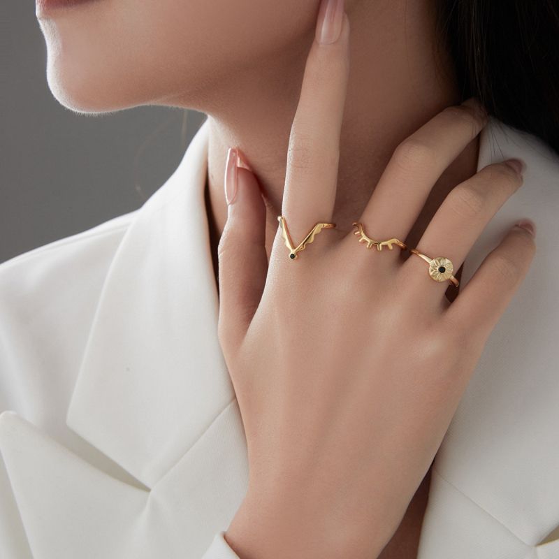 Gros plan d'une main portant trois bagues plaquées or : une bague en forme de V avec une petite pierre noire, une bague avec des motifs d'yeux stylisés, et une bague œil du mal avec une pierre noire centrale.