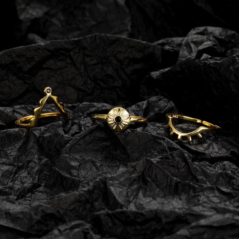 Trois bagues en argent plaqué or sur fond noir : une bague forme triangle avec une pierre noire, une bague marguerite avec une pierre noire au centre, et une bague œil stylisé.