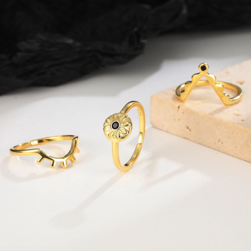Trois bagues dorées sur un fond blanc : une bague œil stylisé, une bague fleur avec une pierre noire au centre, et une bague en forme de couronne avec une pierre noire