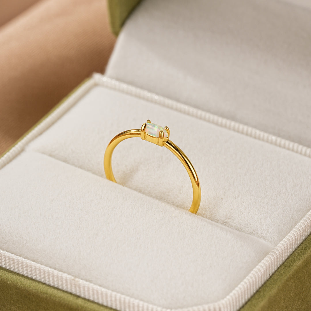 Bague fine en argent plaqué or avec une opale rectangulaire blanche au centre, présentée dans une boîte à bijoux verte.