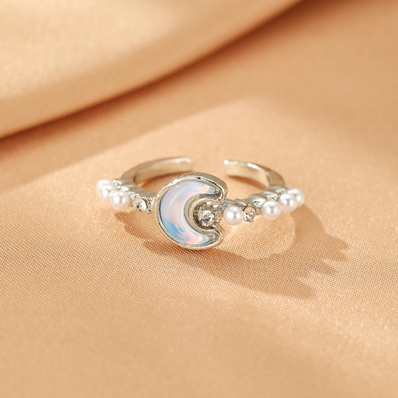 Bague ouverte argentée ornée d'une lune en opale irisée, entourée de petites perles et de cristaux scintillants.