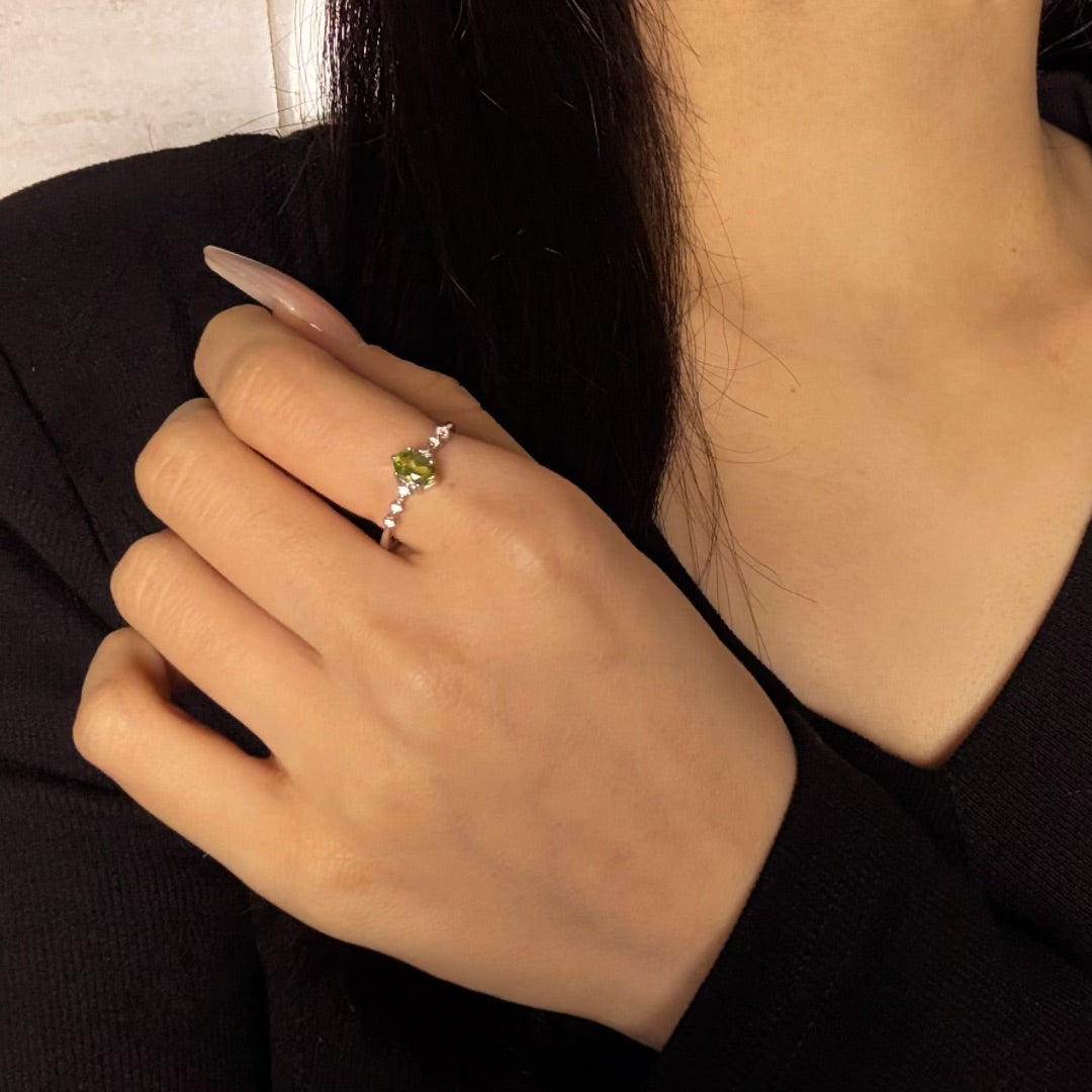 Bague en argent avec une pierre péridot verte en forme de cœur, sertie sur un anneau orné de petites pierres scintillantes, portée à l'annulaire d'une main aux ongles longs et soignés.