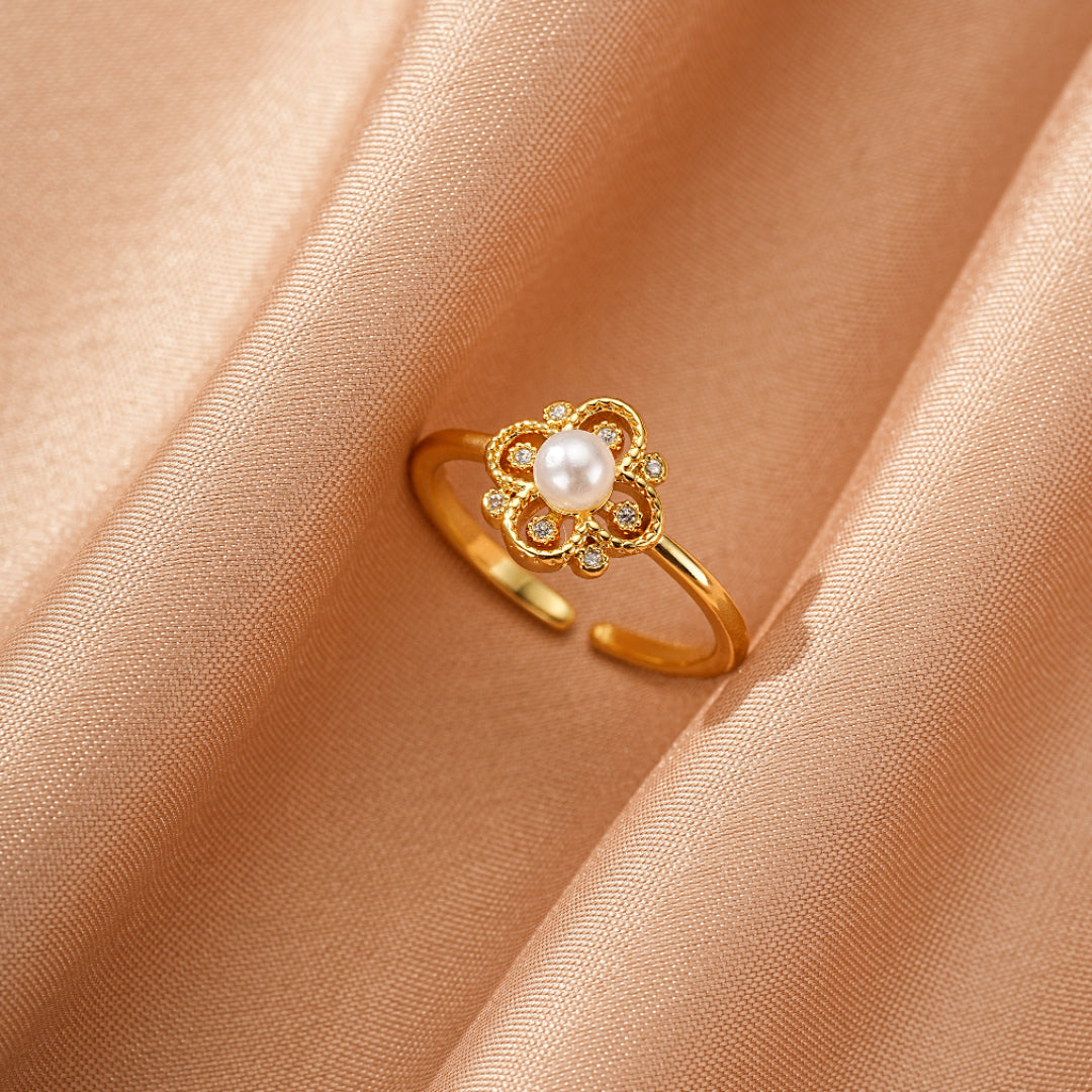 Bague ajustable en métal doré avec un motif de fleur sertie d'une perle blanche et de petits cristaux, posée sur un tissu satiné couleur pêche.