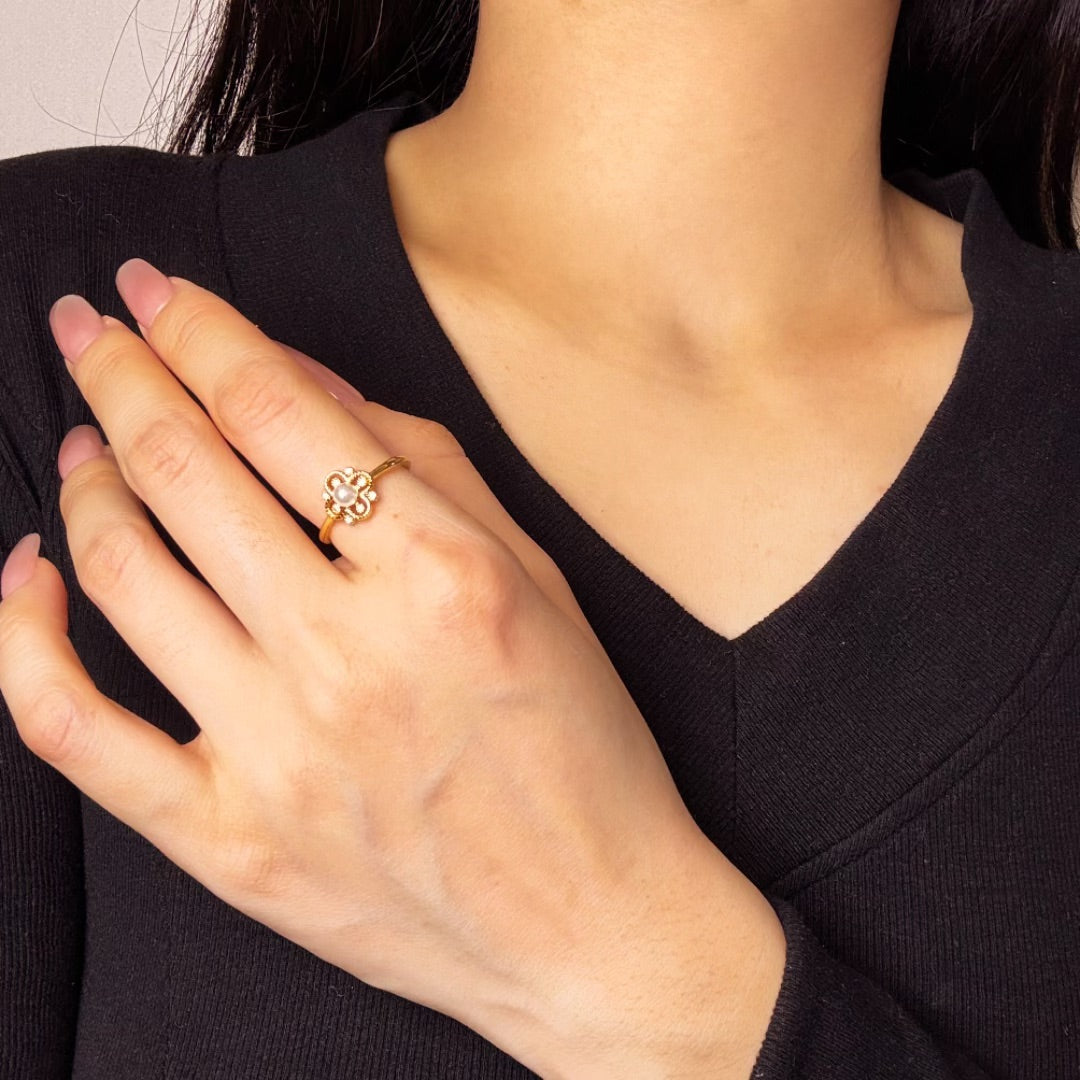 Bague en or jaune avec un motif de fleur stylisée ornée d'une petite perle au centre, portée sur l'annulaire d'une main féminine.