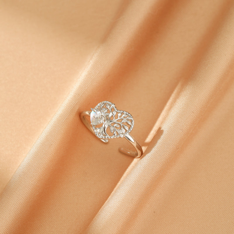 Bague en argent 925 avec un cœur ajouré orné d'un arbre de vie et d'une pierre de lune, posée sur un tissu satiné couleur pêche.