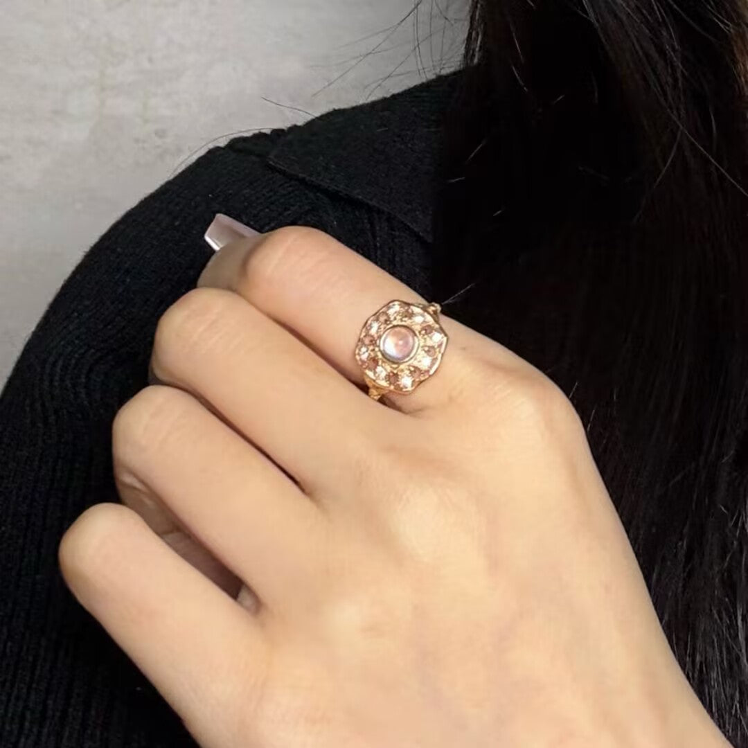 Bague en argent plaqué or avec une pierre de lune centrale entourée de petites pierres, design floral, portée à la main.
