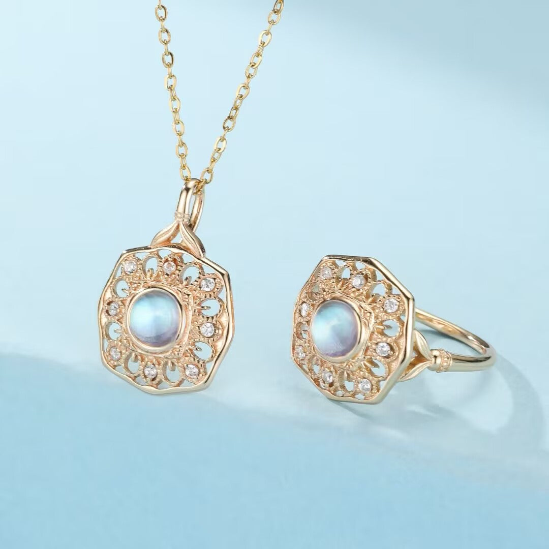 Bague et pendentif en pierre de lune, sertis dans de l'argent plaqué or avec des détails complexes et des zircons cubiques, sur fond bleu clair.
