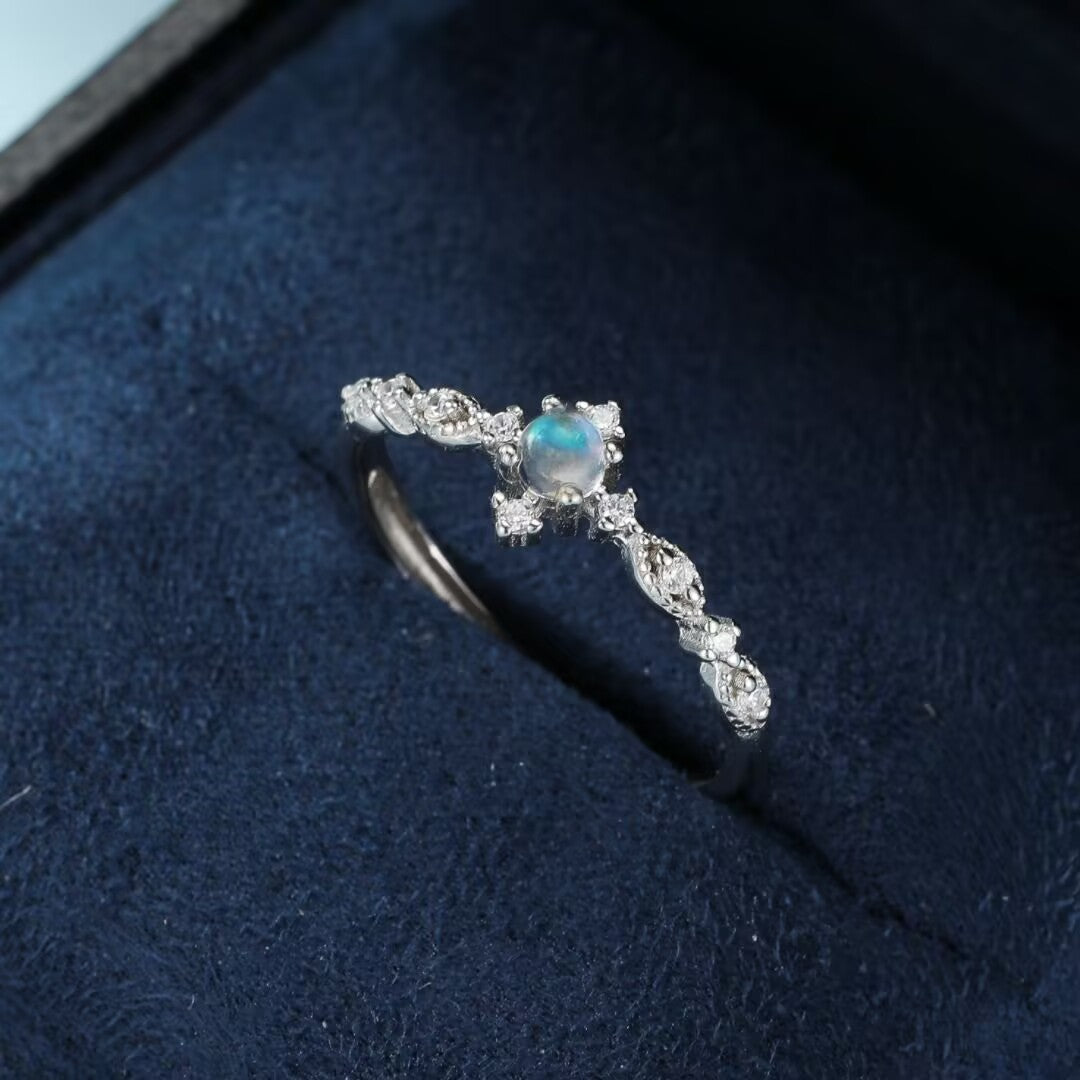 Bague délicate en argent ornée d'une pierre de lune centrale bleutée et de petits diamants scintillants sur le corps de la bague, le tout dans un écrin bleu.