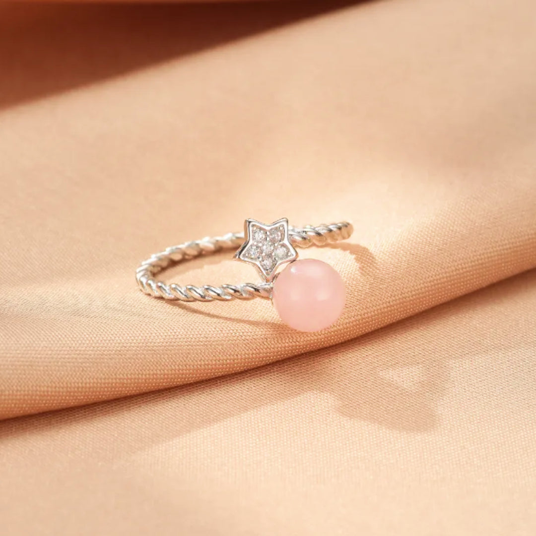 Bague en argent ornée d'une étoile sertie de cristaux et d'une perle ronde en quartz rose sur un fond satiné beige.