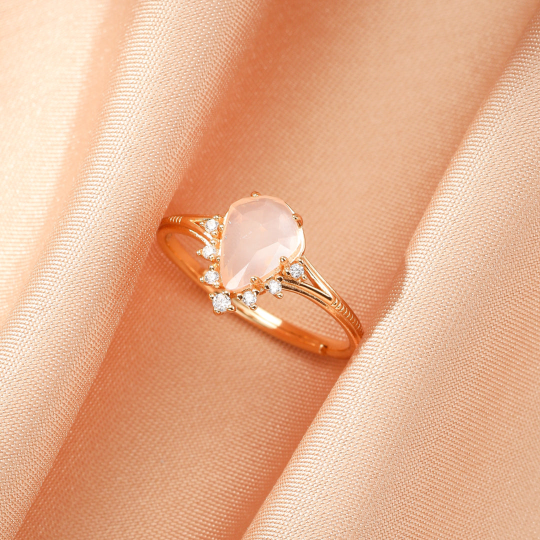 Bague en argent plaqué or 9K avec une pierre de quartz rose en forme de goutte entourée de diamants scintillants et d'une monture élégante, posée sur un fond de soie rose.