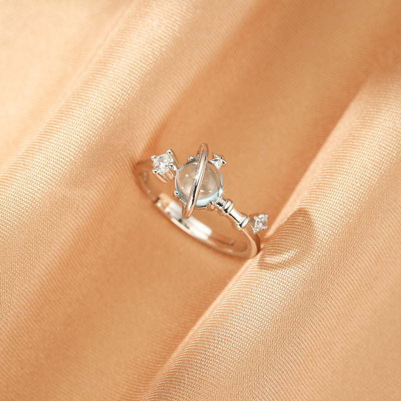 Bague ajustable en argent 925 avec une topaze bleue centrale rappelant une planète, entourée de deux anneaux et ornée de deux petits zircons cubiques, sur un fond satiné couleur pêche.
