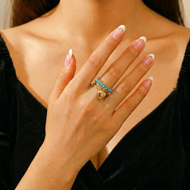 Bague fine dorée avec deux anneaux superposés, ornés de petites perles turquoise et d'un pendentif en forme de cœur doré.