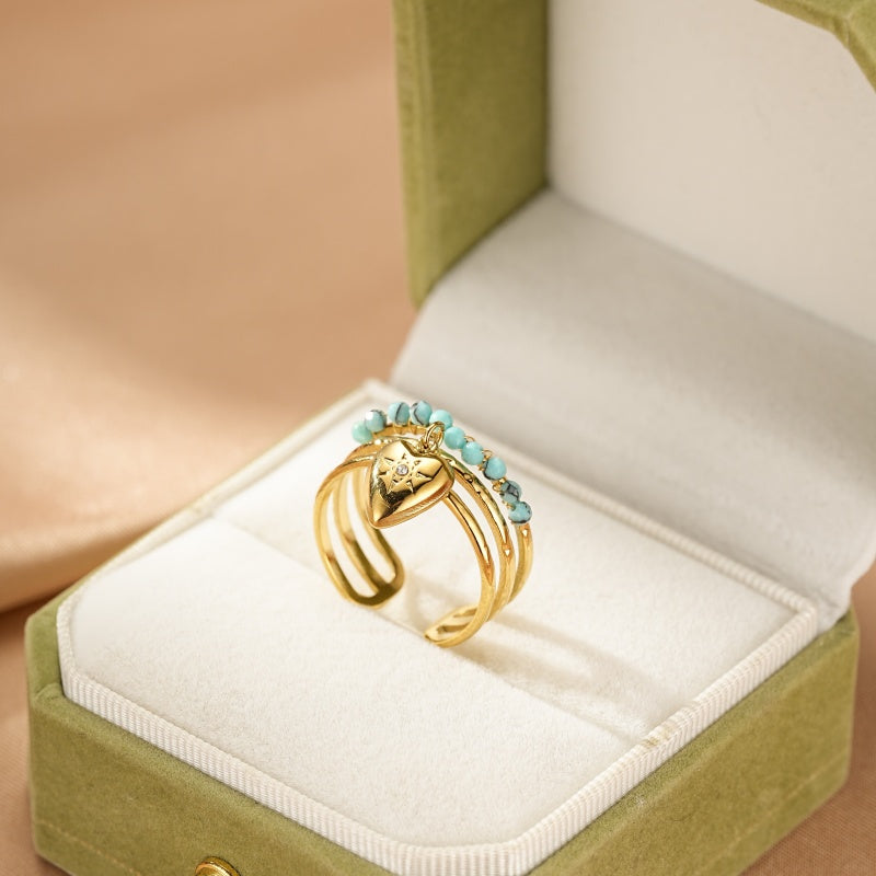 Bague dorée à trois anneaux avec un pendentif en forme de cœur orné d'une petite étoile et de petites perles bleues turquoise sur le dessus, posée dans une boîte à bijoux verte.