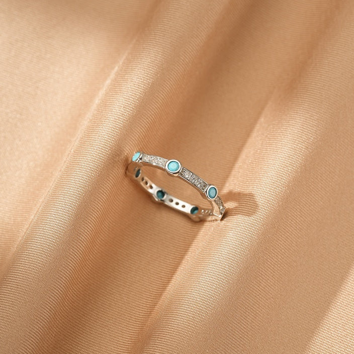 Bague fine en argent 925 sertie de petites pierres turquoise rondes et de cristaux scintillants, posée sur un tissu satiné couleur sable.