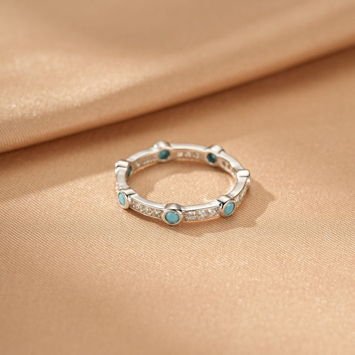 Bague fine en argent ornée de petites pierres turquoise rondes et d'éclats de cristaux, posée sur un tissu satiné beige.
