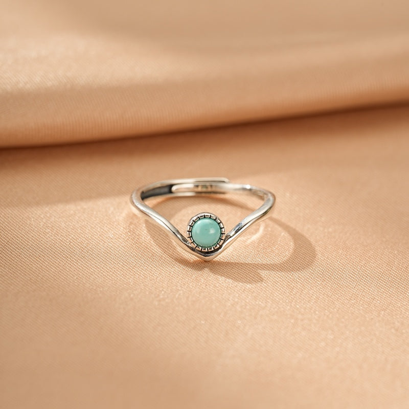 Bague en argent avec une petite turquoise ronde sertie sur une monture en V sur un fond beige satiné.