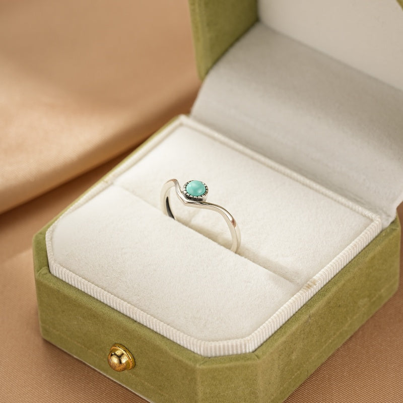 Bague en argent avec une petite turquoise ronde sertie sur une monture ondulée en forme de V, présentée dans un écrin vert.