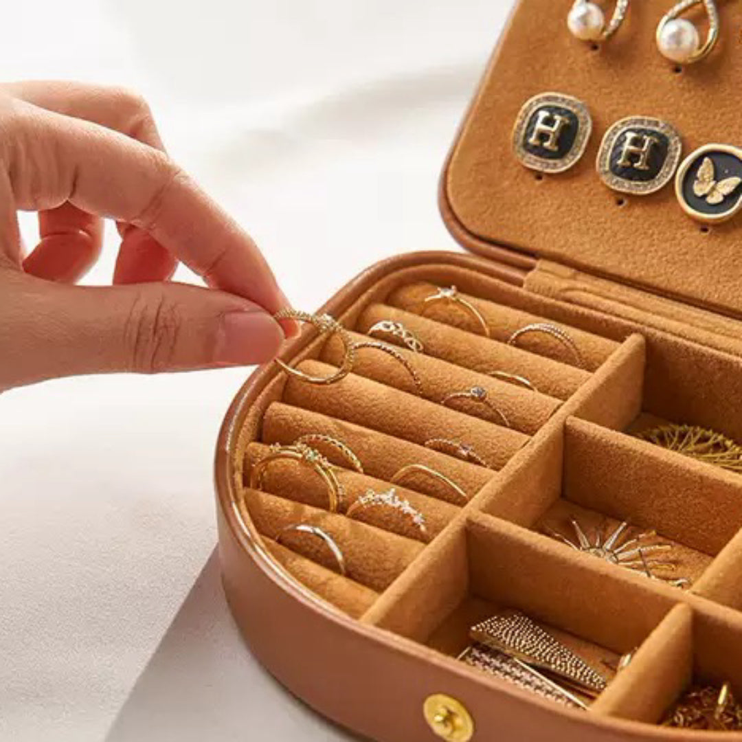 Une main sort une bague d'une boîte à bijoux en cuir marron avec un intérieur en velours brun, organisée avec plusieurs compartiments pour bagues et boucles d'oreilles.