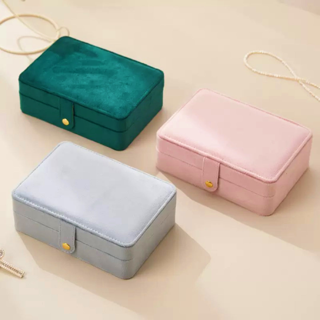 Trois boîtes à bijoux compactes en velours de couleur vert émeraude, rose poudré et bleu ciel, posées côte à côte sur une surface beige.