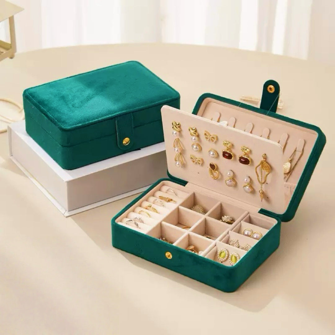 Boîte à bijoux en velours vert émeraude ouverte, révélant des compartiments remplis de bagues, boucles d'oreilles et colliers. Une boîte fermée est visible en arrière-plan.