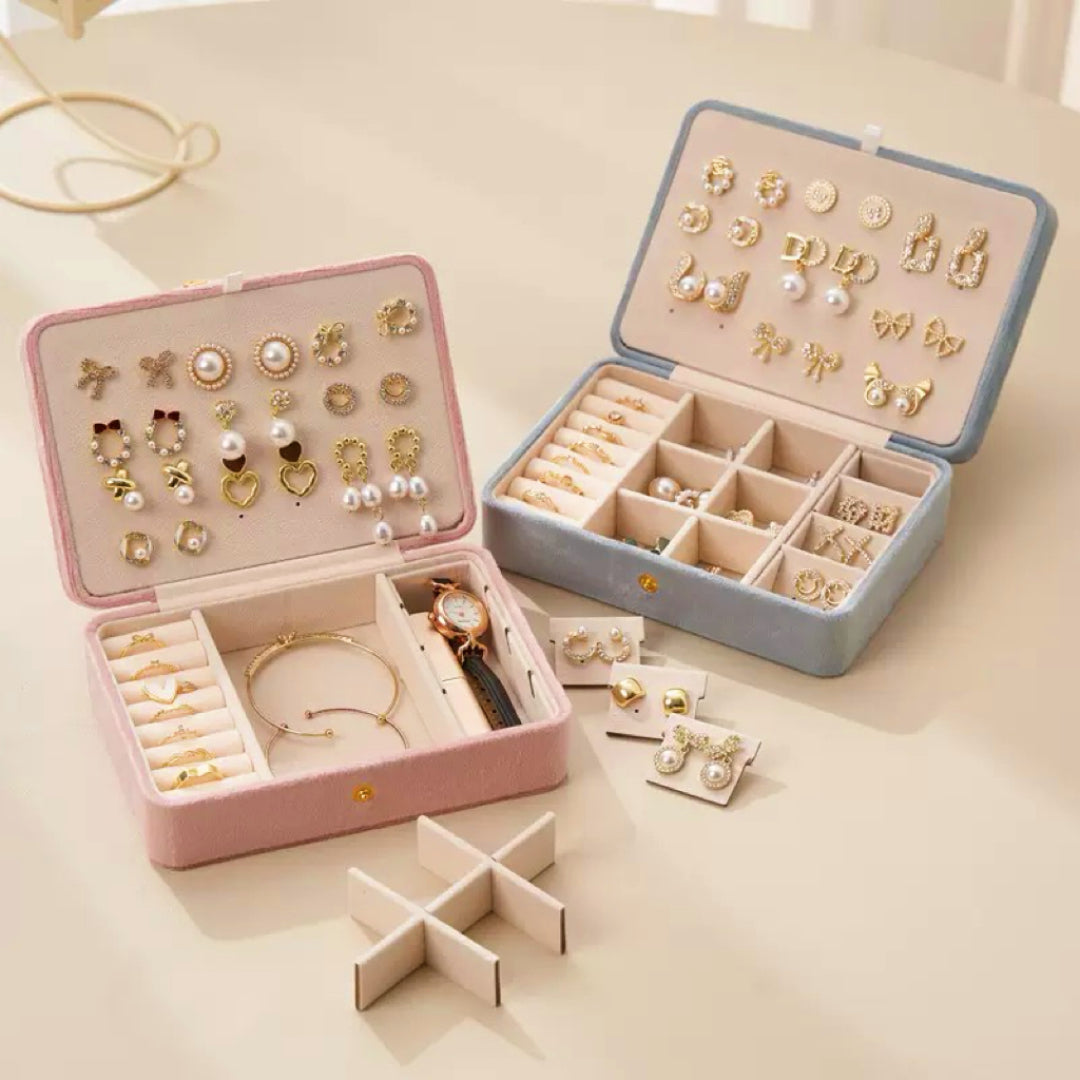 Boîte à bijoux en velours rose et bleue ouverte, remplie de boucles d'oreilles, de bagues et d'une montre, avec des présentoirs à bijoux supplémentaires à proximité.