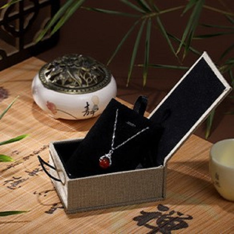 Boîte cadeau zen en lin beige ouverte, présentant un collier délicat avec un pendentif en pierre rouge, sur un fond de bambou et d'un brûle-parfum.