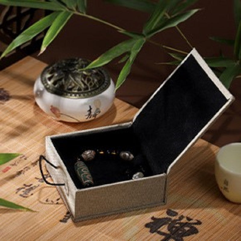 Boîte cadeau zen en lin beige ouverte, contenant un bracelet orné de perles et d'une amulette bouddhiste. Un brûle-parfum en céramique et une petite tasse complètent la scène.